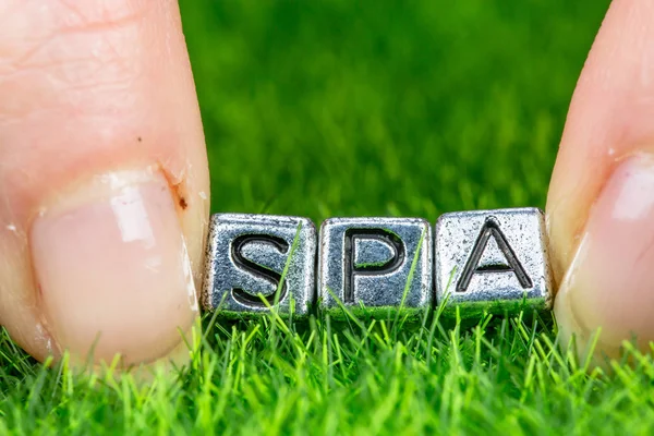 Word Spa skrivet i metall bokstäver som ligger på gräs och hålls mellan fingrarna på en kvinna. Begreppet wellness bakgrund — Stockfoto
