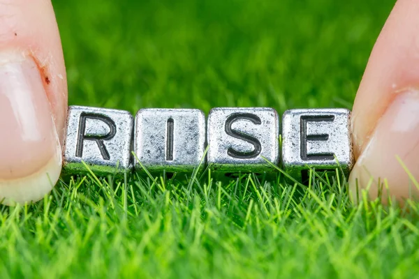 Word RISE written in metal letters laid on grass and held between the fingers of a woman. Concept of hope, goal, overtaking oneself background — Stock Photo, Image