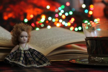 Açık bir kitabın arka planında mini bir bebek, yanan bir şömine, Turki 'den gelen kahve ve Noel ışıkları.