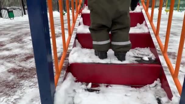 Un niño pequeño en un mono de invierno sube las escaleras a la diapositiva de los niños en invierno — Vídeos de Stock