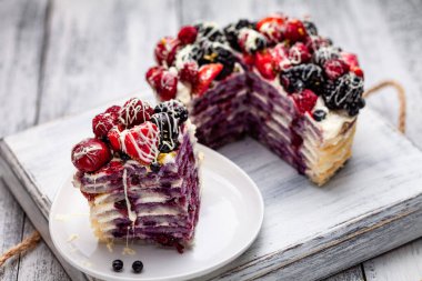a piece of pancake cake with a cap of berries doused with white chocolate on a plate with Belgian muffins and a bottle of syrup on a tray on a white wooden table