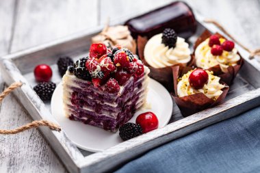 a piece of pancake cake with a cap of berries doused with white chocolate on a plate with Belgian muffins and a bottle of syrup on a tray on a white wooden table