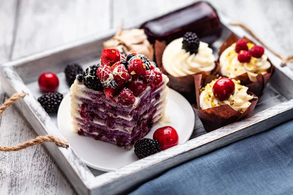 a piece of pancake cake with a cap of berries doused with white chocolate on a plate with Belgian muffins and a bottle of syrup on a tray on a white wooden table