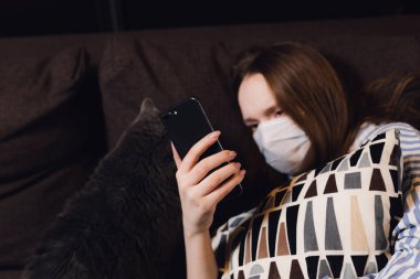 Karantinaya alınmış kız. Kanepede yatarken akıllı telefondan haber okumak. Koronovirüs korunması. Biyolojik koruma salgını salgını konsepti bilim adamı. COVID-19