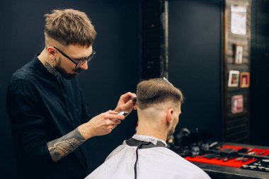 Saç kesimini mükemmel gösteriyor. Berber dükkanında sandalyede otururken genç sakallı bir adam kuaföre tıraş oluyor. dövme