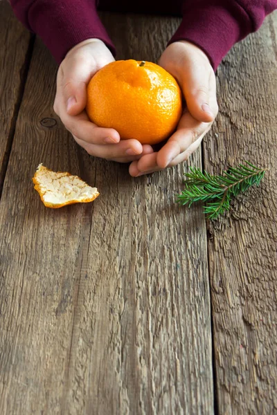 Tangerine and fir branches in children hands close up over rustic wooden background - smell of Christmas time and winter holidays at home concept