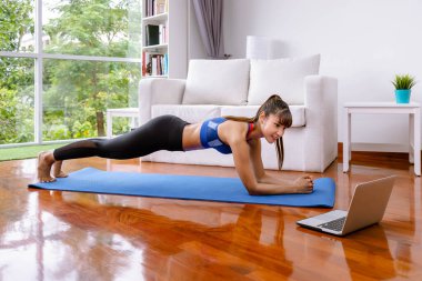 Spor giyimli çekici Asyalı kadın yoga yapıyor ve laptopta özel ders izliyor. Evde oturma odasında tahta egzersizi yapıyor.