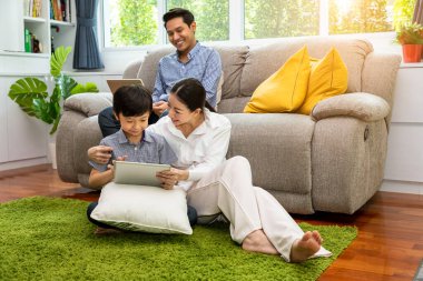 Asyalı çocuk annesiyle tablet kullanırken, baba kanepede oturmuş dizüstü bilgisayarla oturma odasında çalışıyor. Aile ilişkisi kavramı.