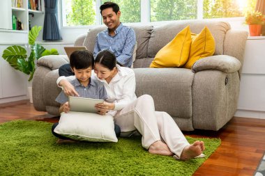 Asyalı bir çocuk annesiyle tablet kullanırken, baba kanepede oturup çalışmak için dizüstü bilgisayar kullanırken, anne ekranı işaret ediyor.
