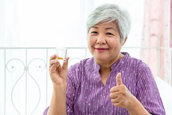 Senior elder woman female looking at measuring glass cup checking medicine, with thumb-up