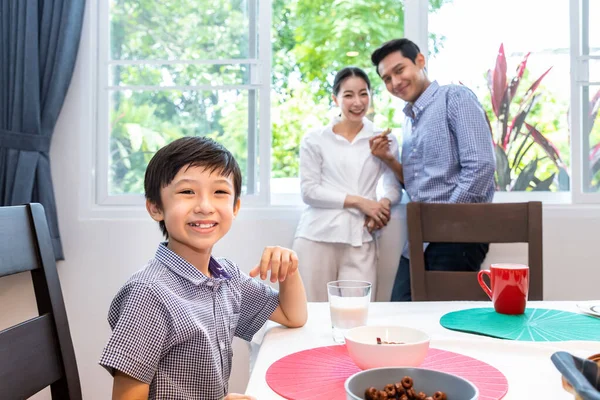 ダイニングルームで幸せなアジアの家族 男の子はダイニングテーブルに座って 笑顔とカメラを見て 両親の父親と母親が背景に立っている — ストック写真