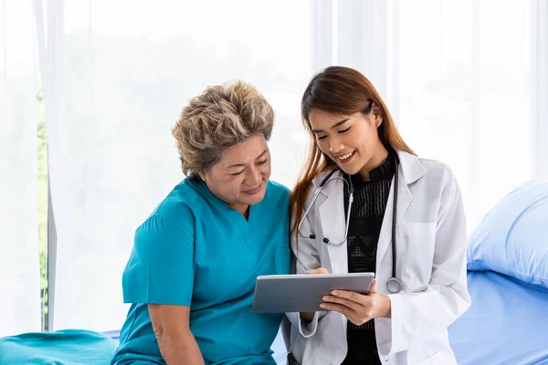 Asian medical doctor take care and explain senior elderly woman female patient sitting on bed with tablet