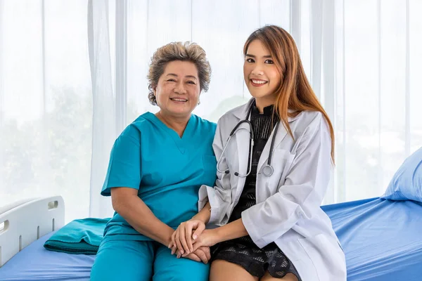 Asian medical doctor holding senior elderly woman female patient hand sitting on bed to comfort and motivate, looking at camera