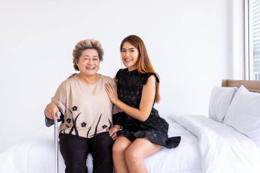 Happy senior elderly Asian woman female mother holding cane with daughter sitting on bed in bedroom, looking at camera clipart
