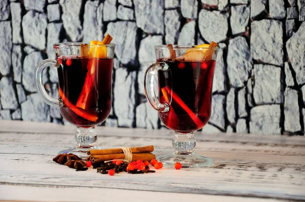 Two glasses of hot wine with spices and slices of fruit stand on a wooden table in a scattering of cinnamon and anise.