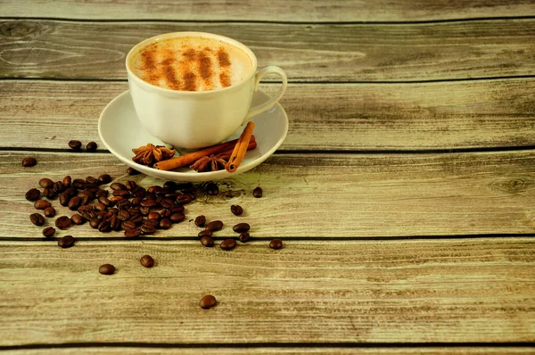 A cup of cappuccino with foam and cinnamon powder stands on a saucer decorated with spice and stands on a wooden table surrounded by coffee beans. Close-up.