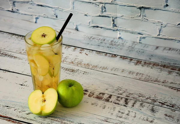 A tall glass of apple juice with ice stands on a wooden table next to green apples. Close-up.