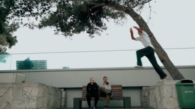 Adam korkuluklardan korkuluklara kadar çok uzun bir atlayış yapıyor. Diğer ikisi izliyor ve peşinden koşuyor. Adam parkour antrenmanı yapıyor. Soğuk bir sonbahar gününde korkuluklara atlıyor. Ters takla atıyor.