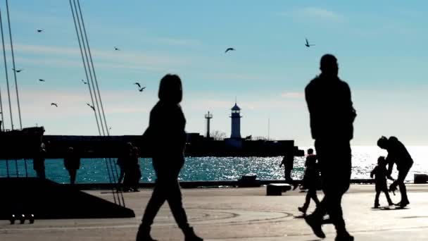 Bright winter sunny day people walk near the sea. Dark silhouettes of people walk on the background of the sea and the lighthouse. The bright sun illuminates the sea, people silhouettes walk, relax in — Stockvideo