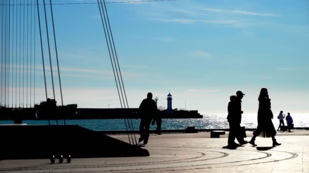 Bright winter sunny day people walk near the sea. Dark silhouettes of people walk on the background of the sea and the lighthouse. The bright sun illuminates the sea, people silhouettes walk, relax in — Stock Video