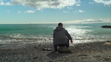 Deniz kenarında bir adam. Bir adam deniz kenarındaki bir kayaya oturur ve güneşli bir kış gününde denize taş atar..