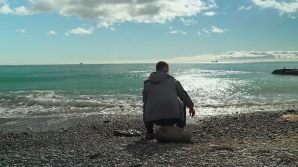 Человек у моря. Человек сидит на камне у моря и бросает камни в море в яркий солнечный зимний день . — стоковое видео