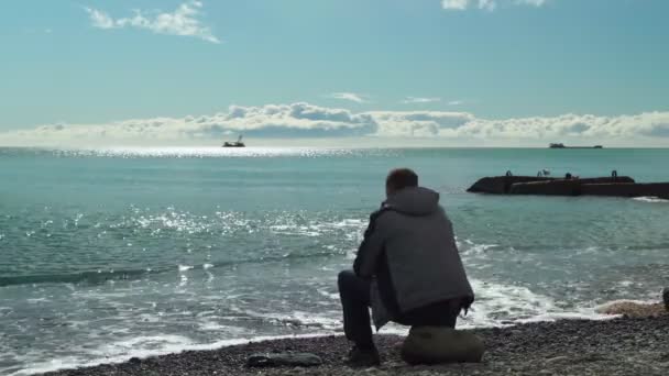 A man by the sea. A man sits on a stone by the sea and throws stones at sea on a bright sunny winter day. — Stock Video