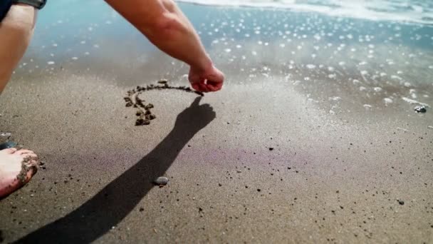 Samiec wyciąga serce przebite strzałą na piasku morskim i fala zmywa rysunek. Malowane serce na piasku morskim, nad brzegiem morza — Wideo stockowe
