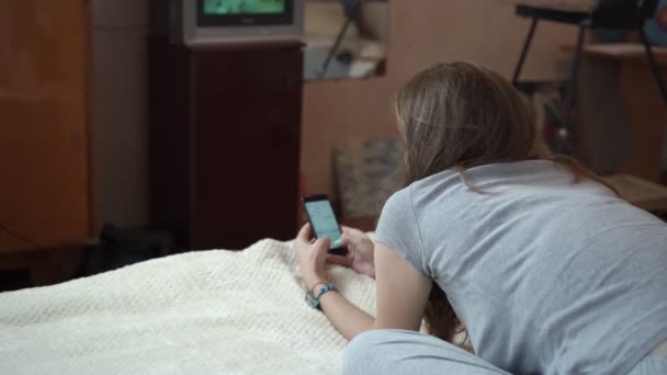 Το κορίτσι ξαπλώνει στο κρεβάτι σε ένα μικρό δωμάτιο και βλέπει μια μικρή τηλεόραση. — Αρχείο Βίντεο