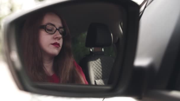 La ragazza in macchina. La ragazza al volante di una macchina si guarda allo specchio laterale e si dipinge le labbra con il rossetto rosso — Video Stock