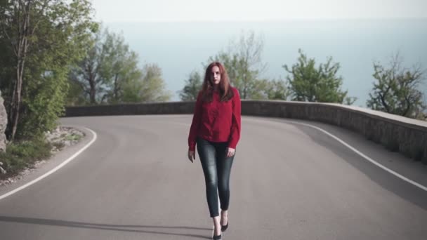 A tall girl in a red shirt and red hair is walking along the dividing line along the center of the road. — Stock Video