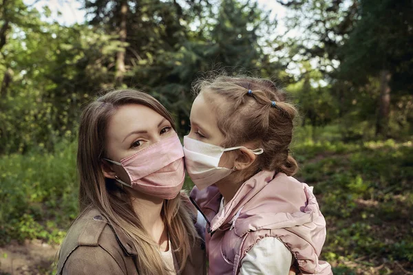 Açık havada tıbbi maske takmış küçük bir kızla annenin portresi. Anne ve kızı koruyucu maskelerle öpüşüyor.