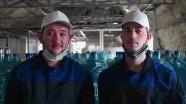 Saf su bitkisi. Özel üniformalı iki çalışan ve koruyucu maske takmış arka planda büyük su şişelerinin yakın plan portresi. 4k