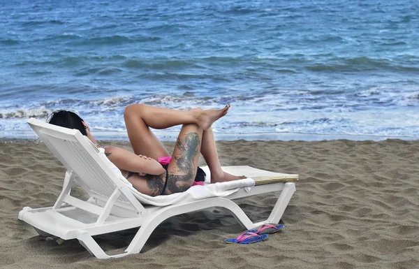 Hermosa, erótica, jovencita en traje de baño, un tatuaje, tomando el sol en la playa tumbada en una tumbona junto al mar — Foto de Stock