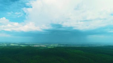 Aerial drone view of big green dense forest growing on hills, beautiful village landscape and blue sky with white clouds.