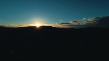 Rising aerial view across populated suburb housing to wide mountain silhouetted by sunrise glow on horizon.