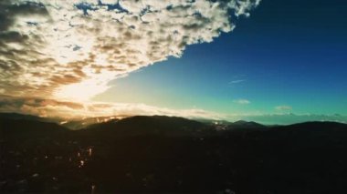 High altitude flight above populated landscape with misty silhouetted mountain range on horizon and view on sunset with fluffy clouds.