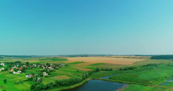 Vista aérea sobre pequeño pueblo con casas en verde valle de campos cultivados y estanques bajo cielo azul . — Vídeo de stock