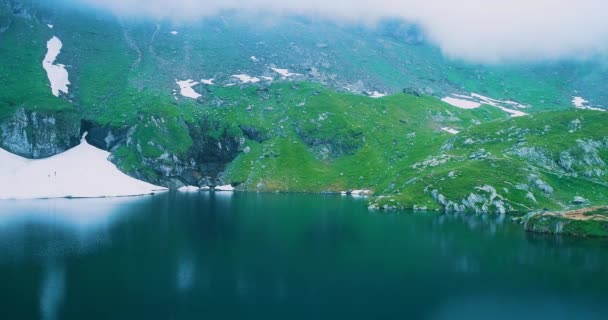 Uitzicht op Transfagarash meer op de bodem van de heuvel met sneeuw en twee toeristen klimmen. — Stockvideo