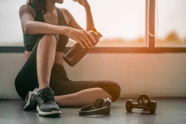 Spor salonunun yanına yerleştirilmiş beyaz bir protein ve halterle egzersiz yapan spor salonundaki kadın. Antrenmandan sonra dinleniyor. Güzel bir genç kadın spor salonunda otururken gözlerini kaçırıyor..