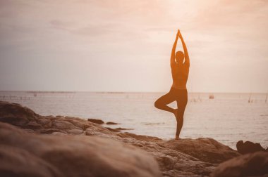 Taze ve sağlıklı olmak için gün batımında deniz manzaralı kayalarda yoga yapan bir kız..