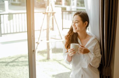 Asya 'nın evinde yaşayan, tatilde yatak odasında bir fincan sıcak kahve içen genç bir kadın. Asya, Asya, rahatla, yalnız, teknoloji, yaşam tarzı konsepti.