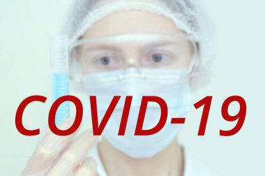 Kadın bir doktor elinde mavi bir sıvıyla bir test tüpü tutuyor. Kırmızı covid-19 yazıtları..