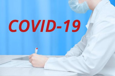 Beyaz önlüklü doktor masada oturuyor ve belgeleri imzalıyor. Kırmızı covid-19 yazıtları..