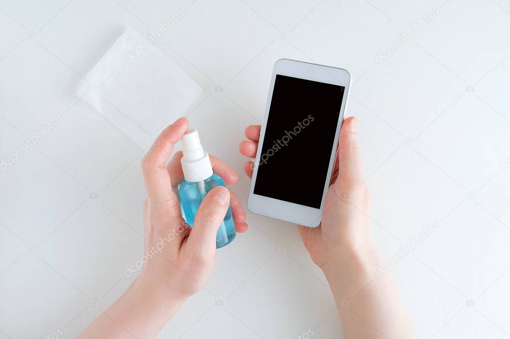 Girl hands sanitizes a white smartphone with a disinfectant in a spray bottle.