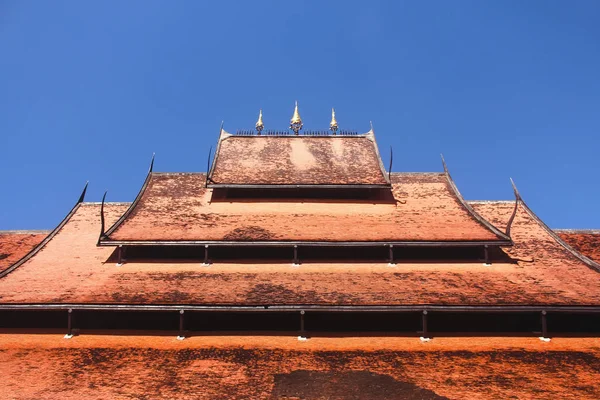 Traditional Thai layered roof with tiered umbrellas at Baandam Museum of Art - Black Temple - against blue sky.
