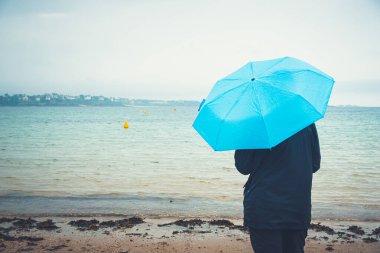 Kasvetli ve yağmurlu bir günde mavi şemsiyeli kadın deniz kıyısında duruyor. Kopyalama alanı olan ayarlanmış resim.