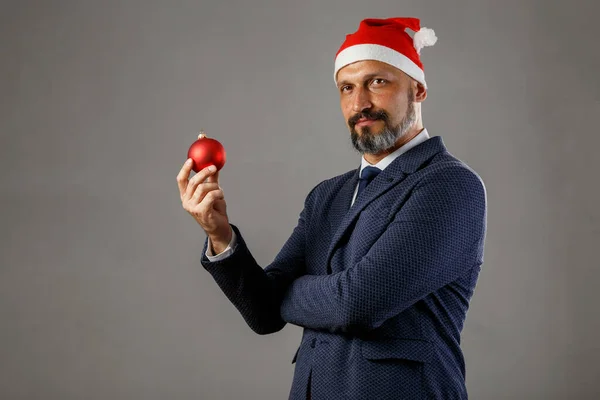 Mavi ceketli sakallı bir adam ve gri arka planda yeni yıl şapkası olan ladin için kırmızı bir Noel topu tutan bir adam. Genç Noel Baba kavramı.