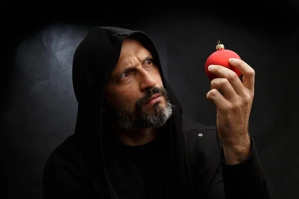 Portrait d'un homme chauve avec une barbe dans une capuche noire avec une boule de Noël rouge sur un fond gris sale. Concept de jeune Père Noël . — Photo