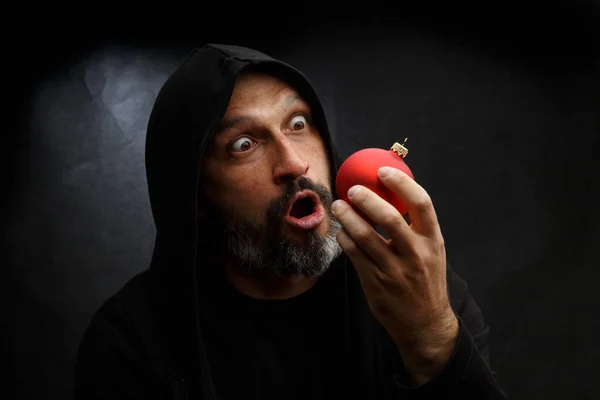 Portrait d'un homme chauve avec une barbe dans une capuche noire avec une boule de Noël rouge sur un fond gris sale. Concept de jeune Père Noël . — Photo